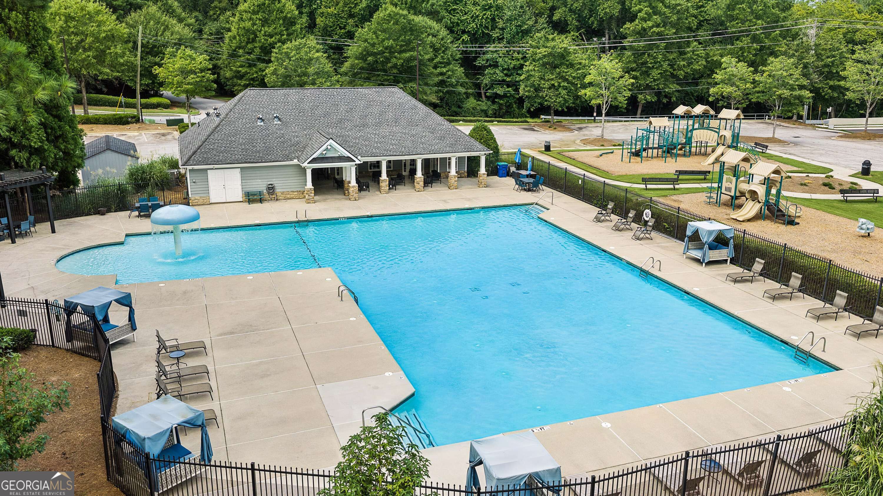 Community pool with a mushroom fountain, fenced deck, covered seating, and a playground beside the clubhouse.