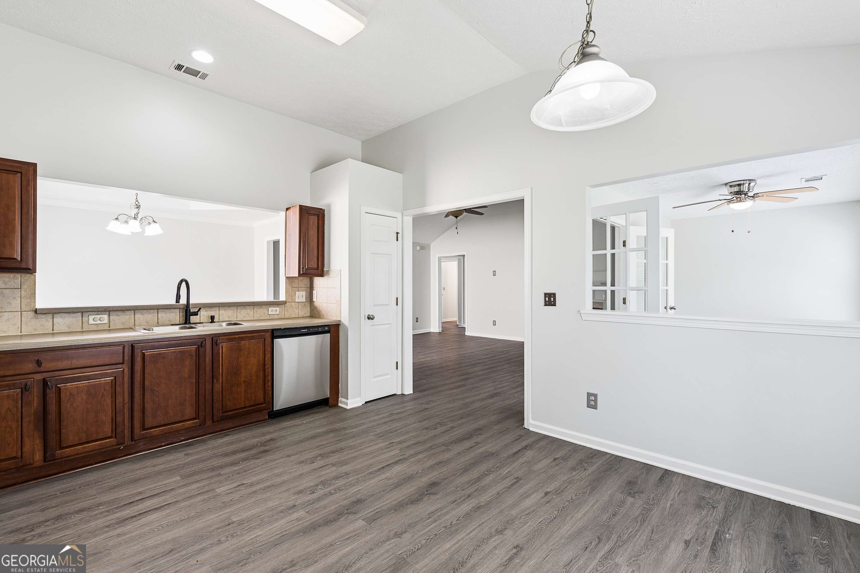 Kitchen with dark wood cabinets and stainless steel dishwasher opening to an empty room with gray flooring.
