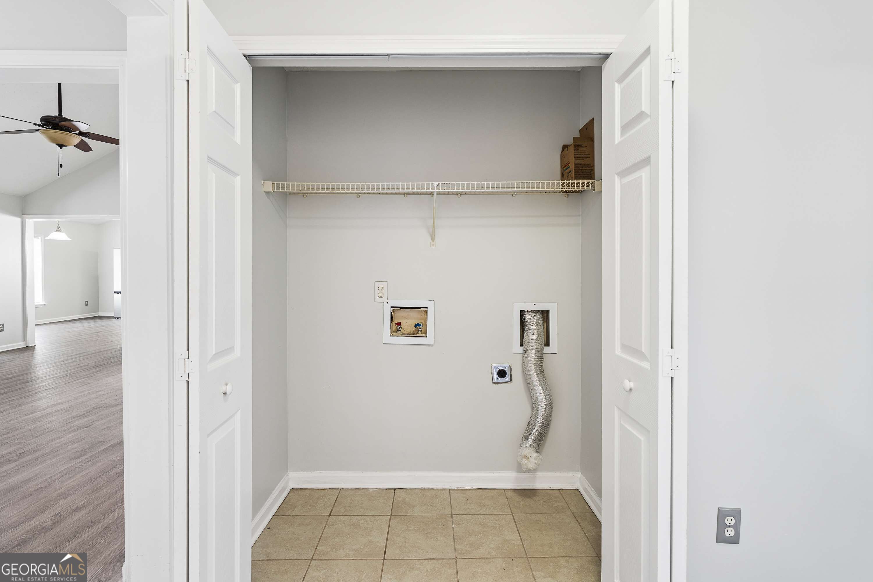 Empty laundry closet with a wire shelf, washer hookups, and a hanging dryer vent hose.