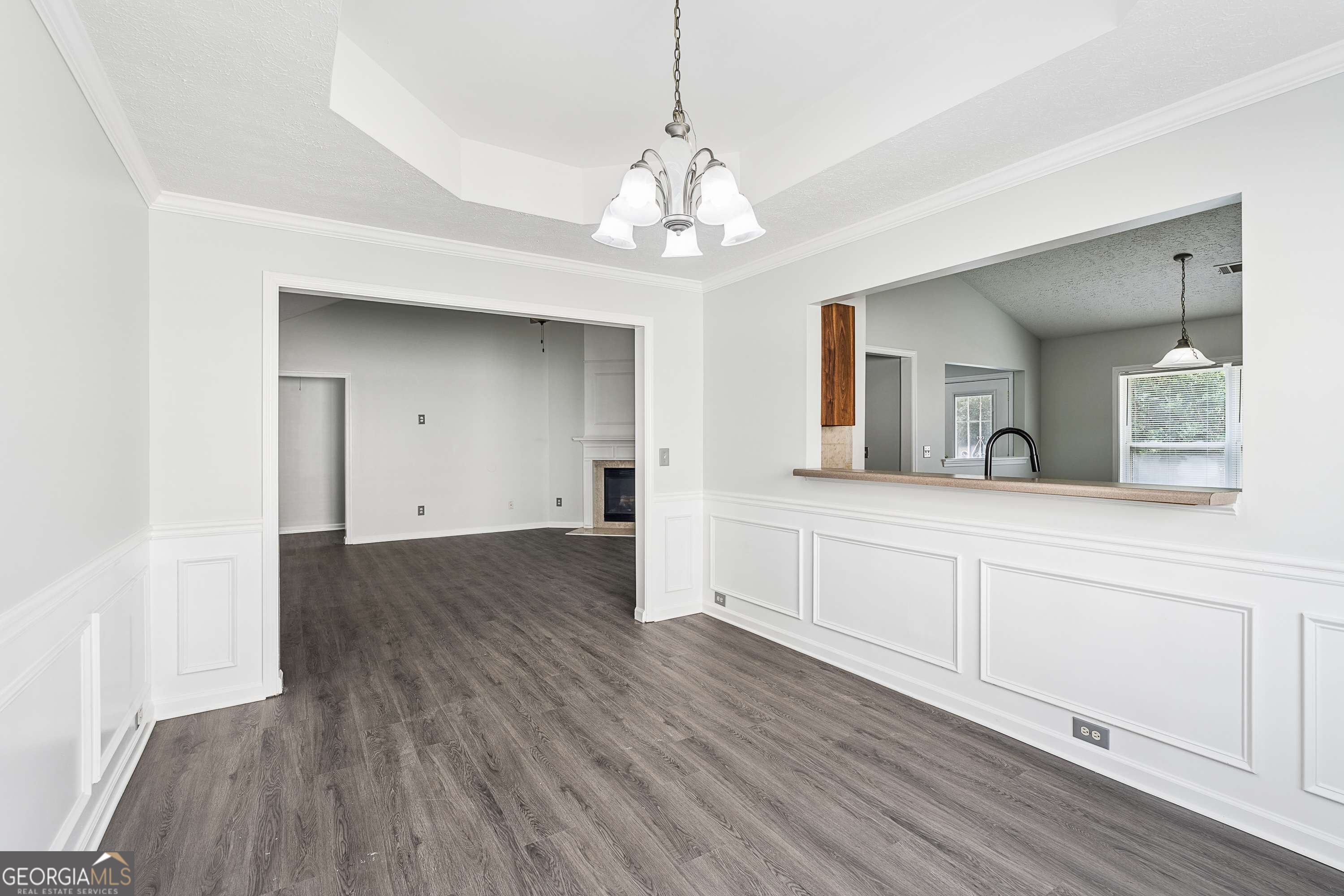 Dining area with dark flooring and white wainscoting, opening to the kitchen and a living room with fireplace.
