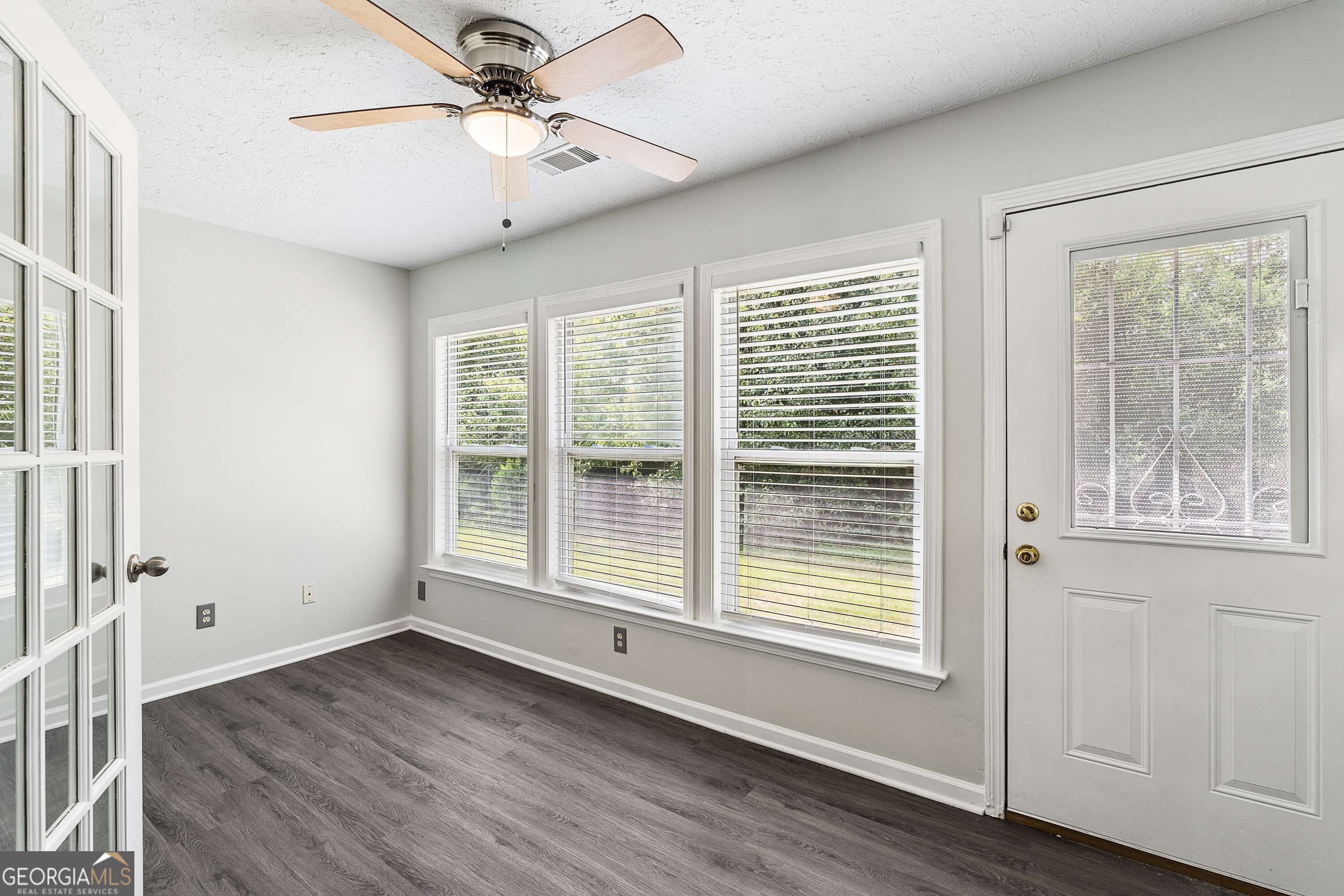 Empty room with dark flooring, a ceiling fan, three blind-covered windows, and a glazed exterior door.