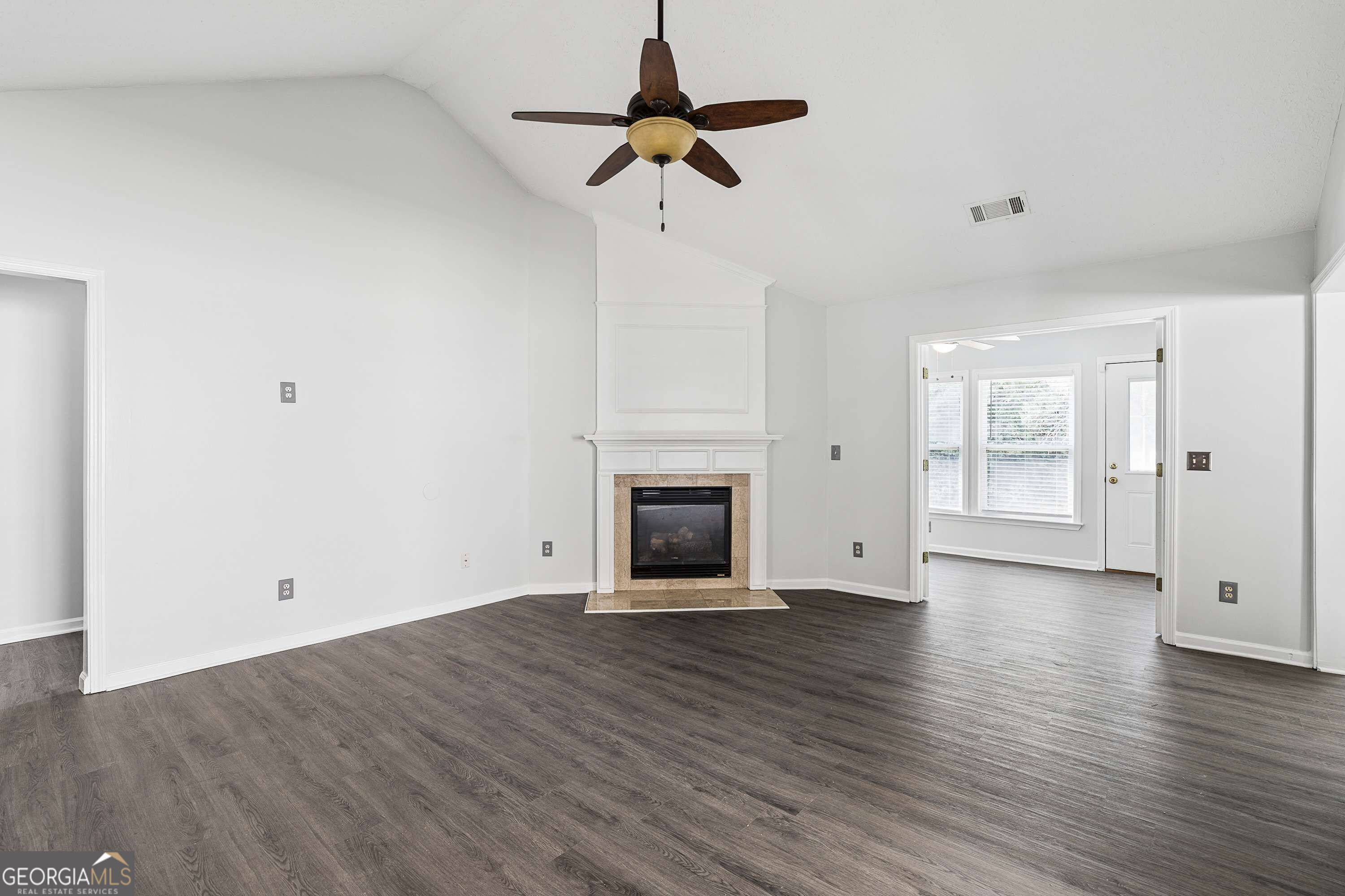 Empty living room with a vaulted ceiling, central fireplace, and dark wood-look flooring.