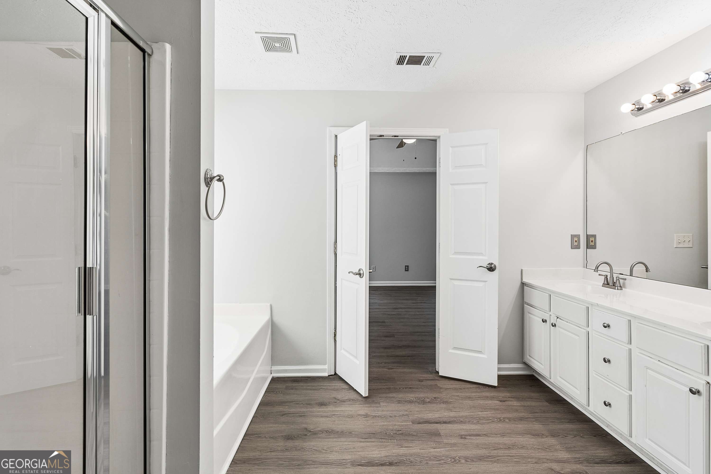 Bathroom with a glass shower, bathtub, and long white vanity beside an open doorway.