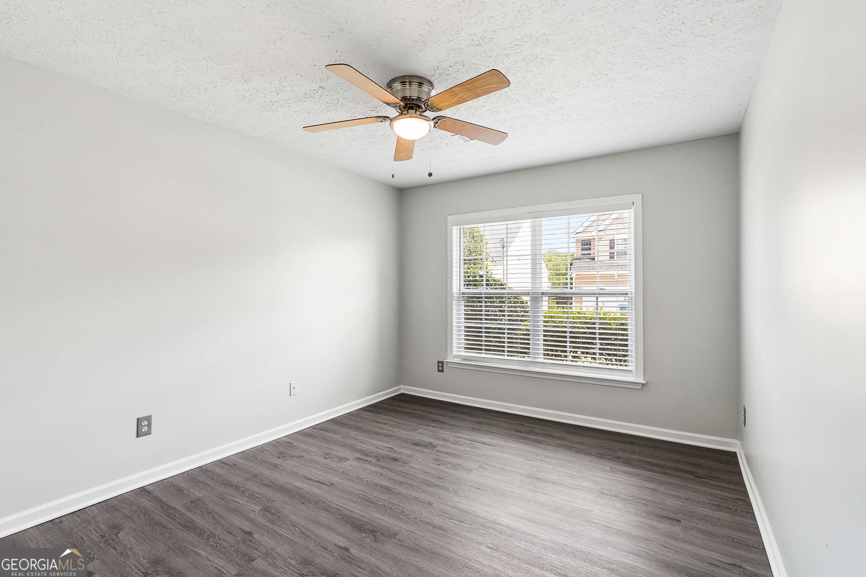 Empty room with gray walls, dark flooring, a large window with blinds, and a ceiling fan.