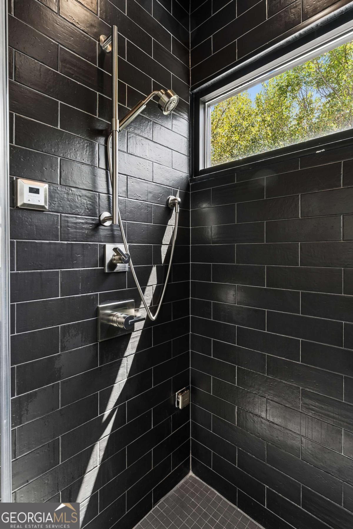 Walk-in shower with dark tile, brushed metal fixtures, and a high window overlooking leafy trees.