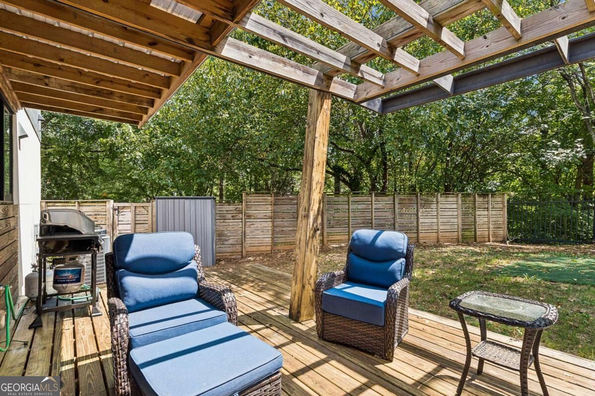 Backyard deck with a timber pergola, blue-cushioned seating, and a grill beside a leafy fenced yard.