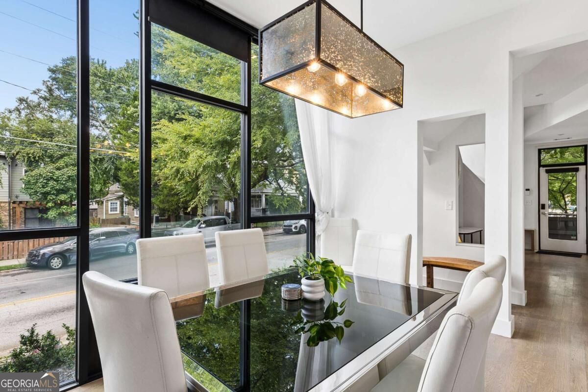 Dining room with a glass-top table, white chairs, and tall windows overlooking a leafy street.