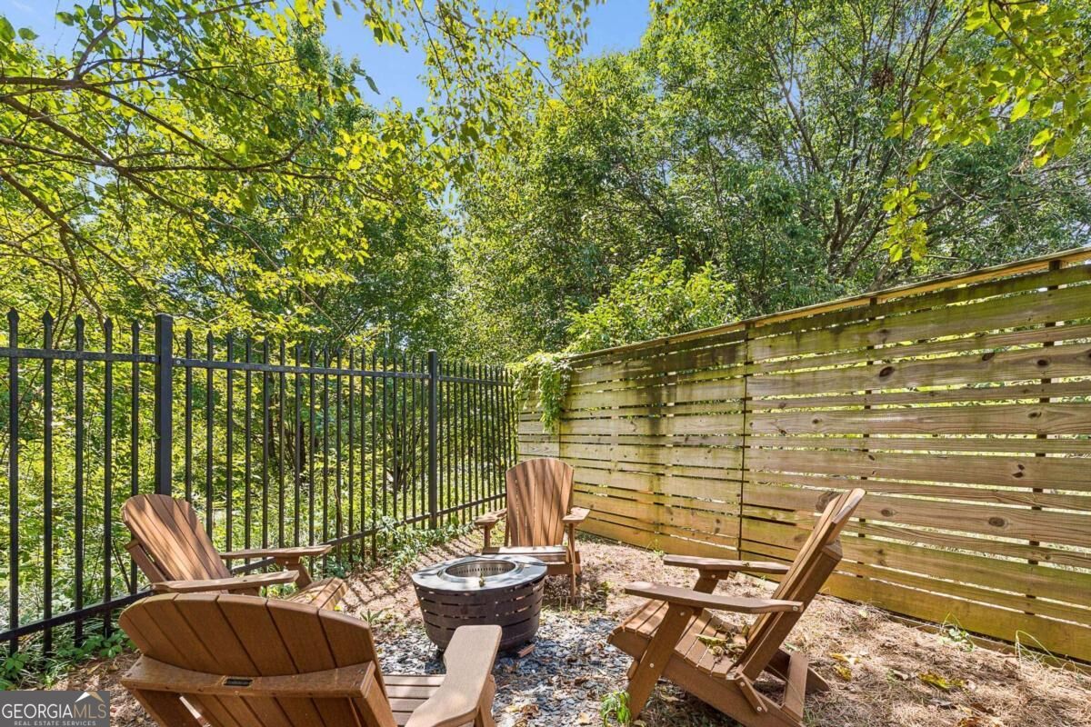 Fenced patio with wooden Adirondack chairs and a central fire pit beneath leafy trees.