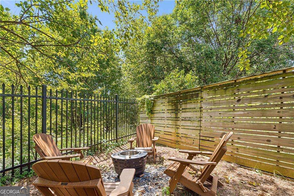 Fenced patio with Adirondack chairs around a fire pit beneath leafy trees.