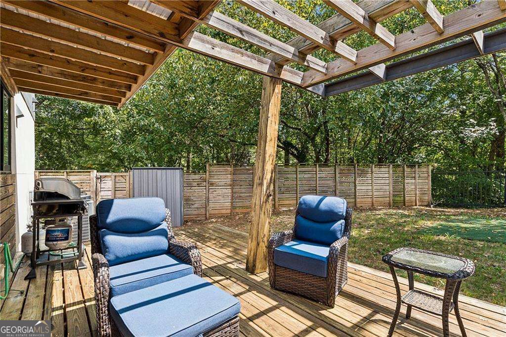 Wooden deck beneath a pergola with blue lounge chairs, a grill, and a fenced wooded yard.