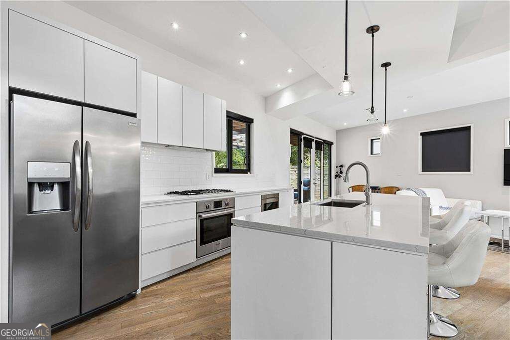 Modern white kitchen with a large island, stainless steel appliances, and an adjoining furnished living area.
