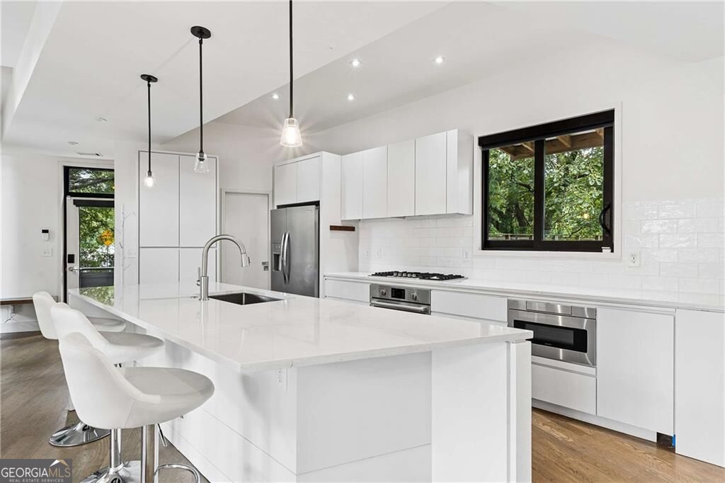 Modern white kitchen with a large island, stainless steel appliances, and a window overlooking trees.