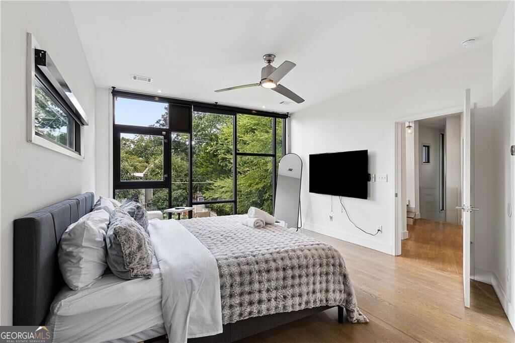Bedroom with a bed beside wide windows overlooking leafy trees and hardwood floors.