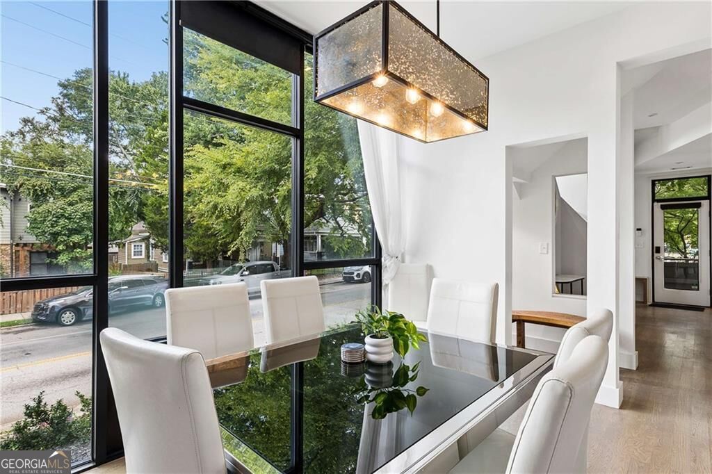 Dining area with a glass table, white chairs, and floor-to-ceiling windows overlooking a leafy street.