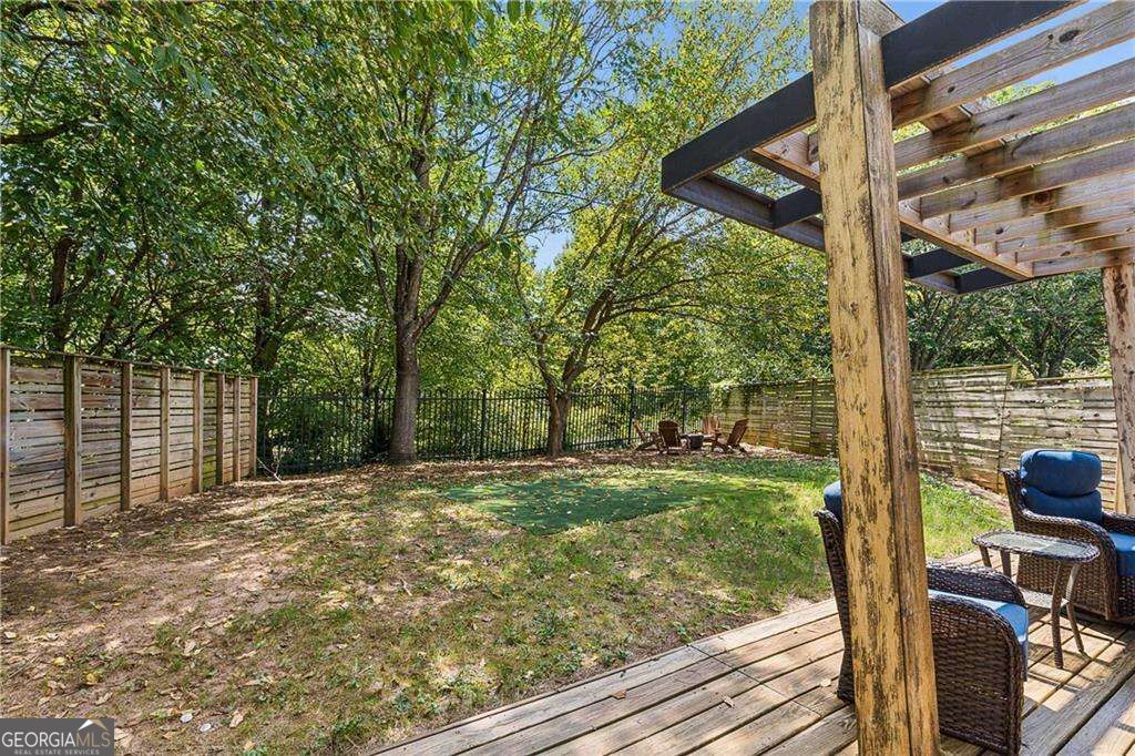 Fenced backyard with a wooden deck, pergola, outdoor seating, and mature trees.