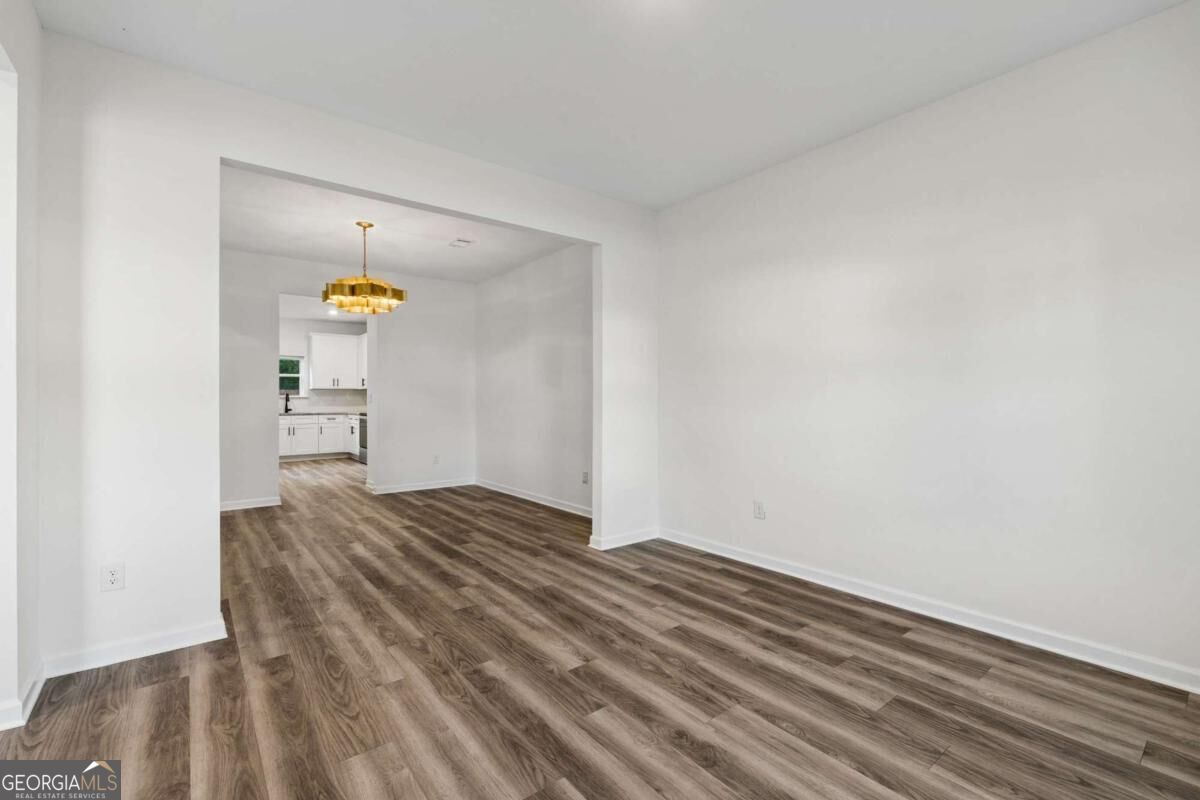 Empty dining area with brown wood-look flooring and a gold chandelier, opening to a white kitchen.