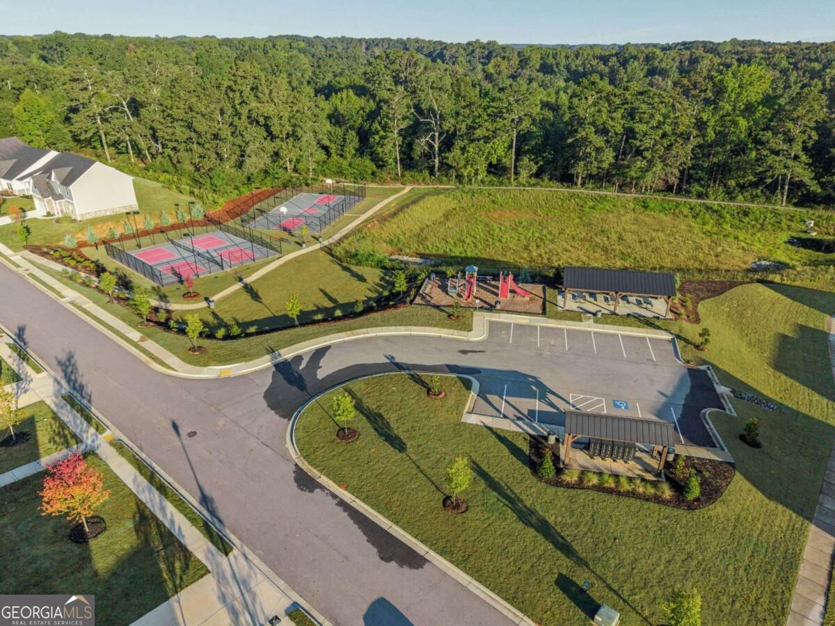 Aerial view of community sport courts and playground beside parking and wooded surroundings.