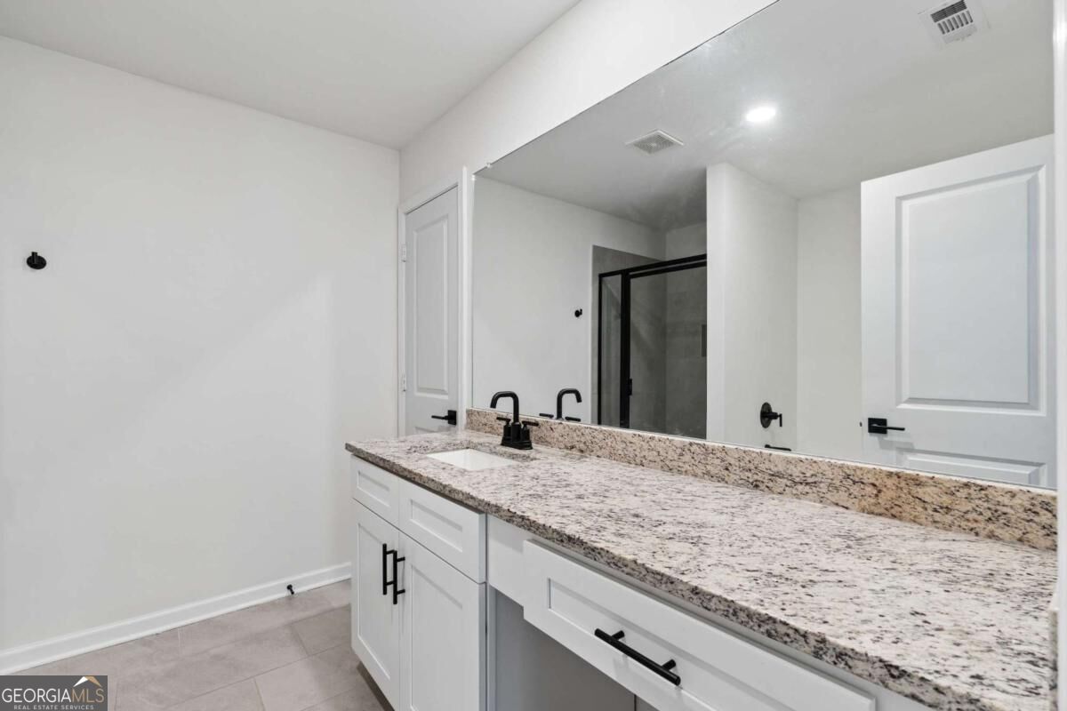 Bathroom vanity with speckled granite countertop, white cabinetry, black faucet, large mirror, and glass shower.