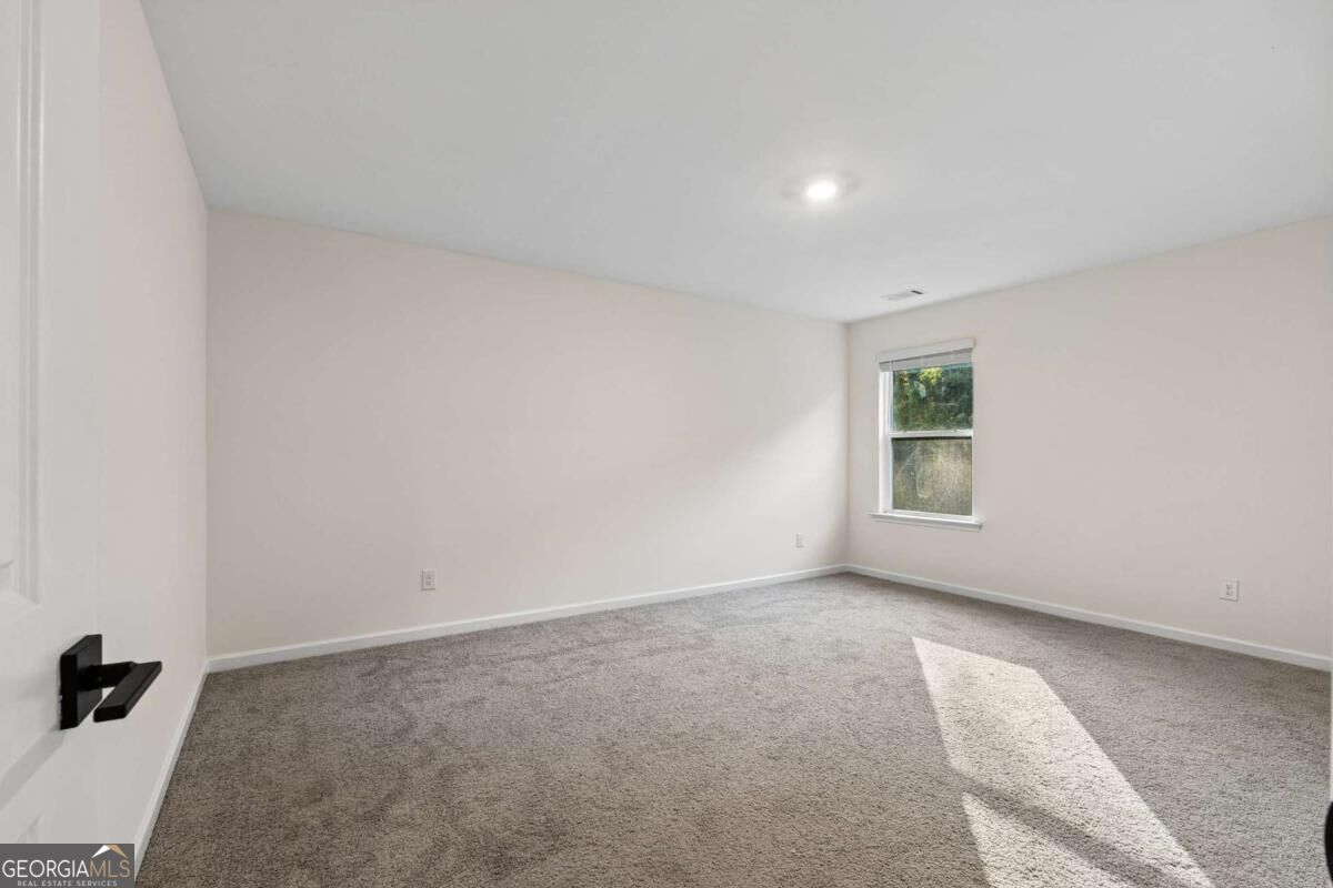 Empty carpeted bedroom with pale walls, a single window, and sunlight across the floor.