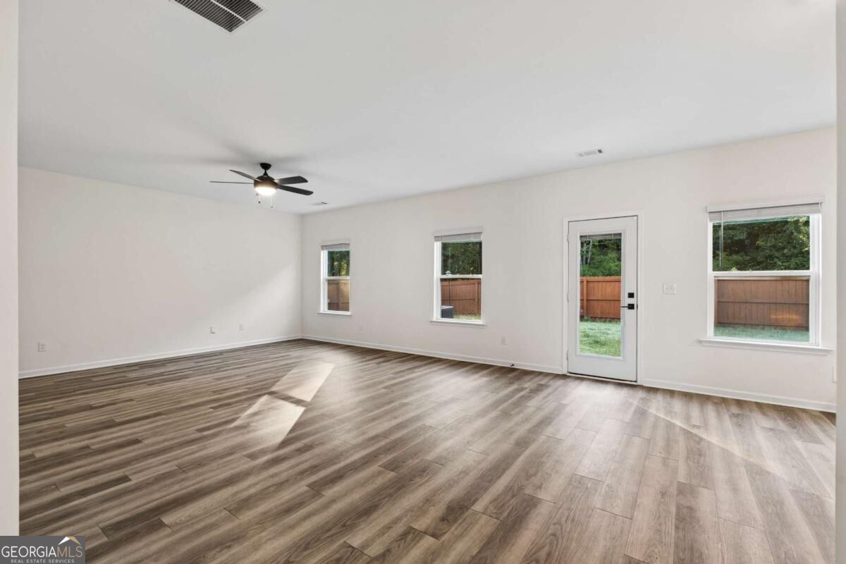 Empty living room with plank flooring, a ceiling fan, and windows overlooking a fenced yard.