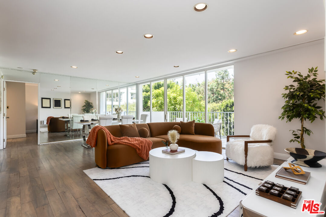 Staged living room with a brown sectional, white coffee tables, and tall windows overlooking trees.