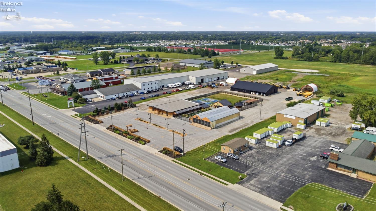 Aerial view of commercial buildings, parking lots, and open fields along a road.