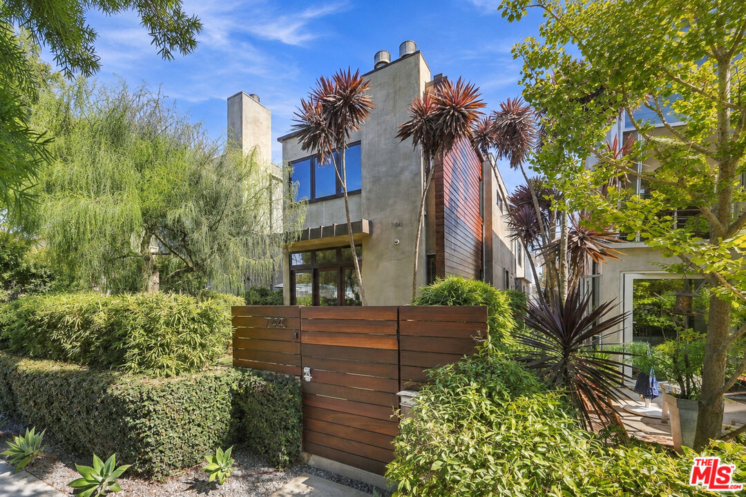Modern concrete-and-wood home behind a broad wooden gate, framed by mature trees and lush landscaping.