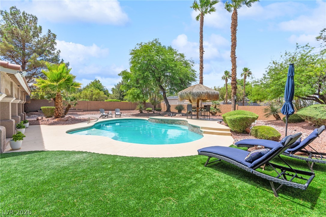 Backyard swimming pool with palm trees, a thatched shade umbrella, lawn, and lounge chairs.