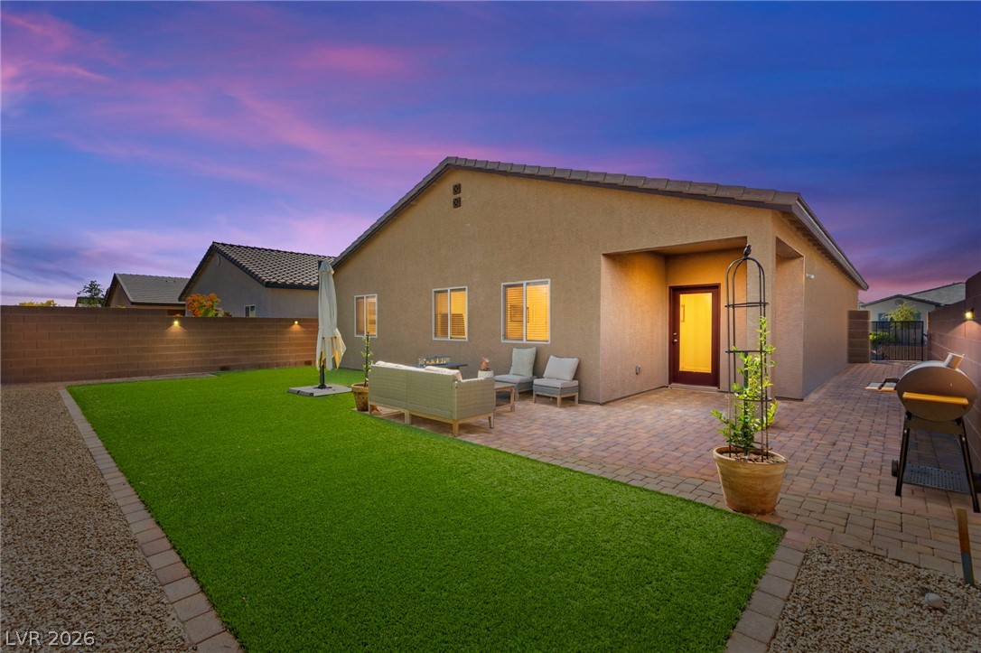 Backyard patio with outdoor seating, artificial lawn, and a glowing entry at dusk.