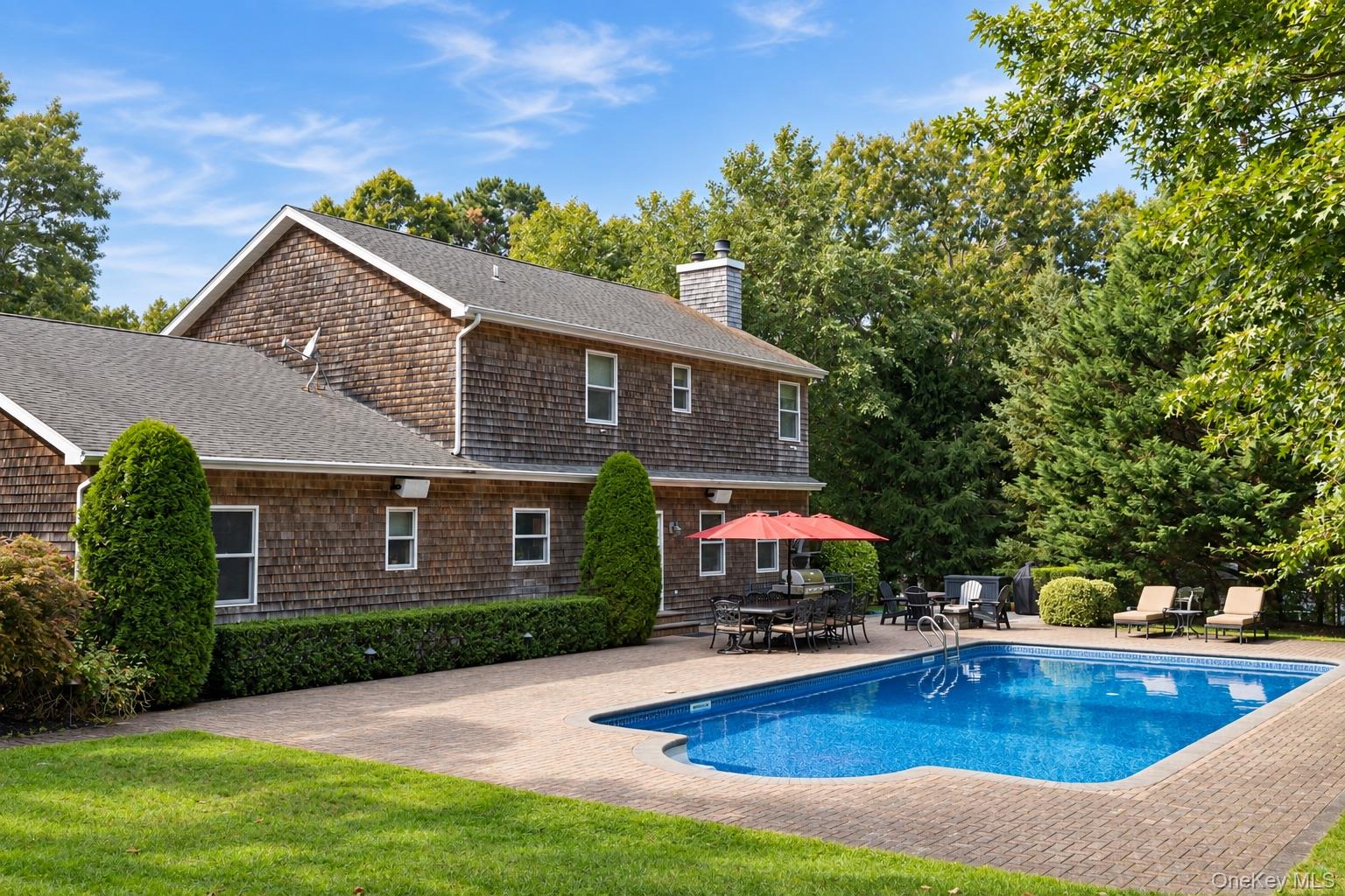 Shingle-sided house beside a backyard pool, with a paved patio and red umbrella.
