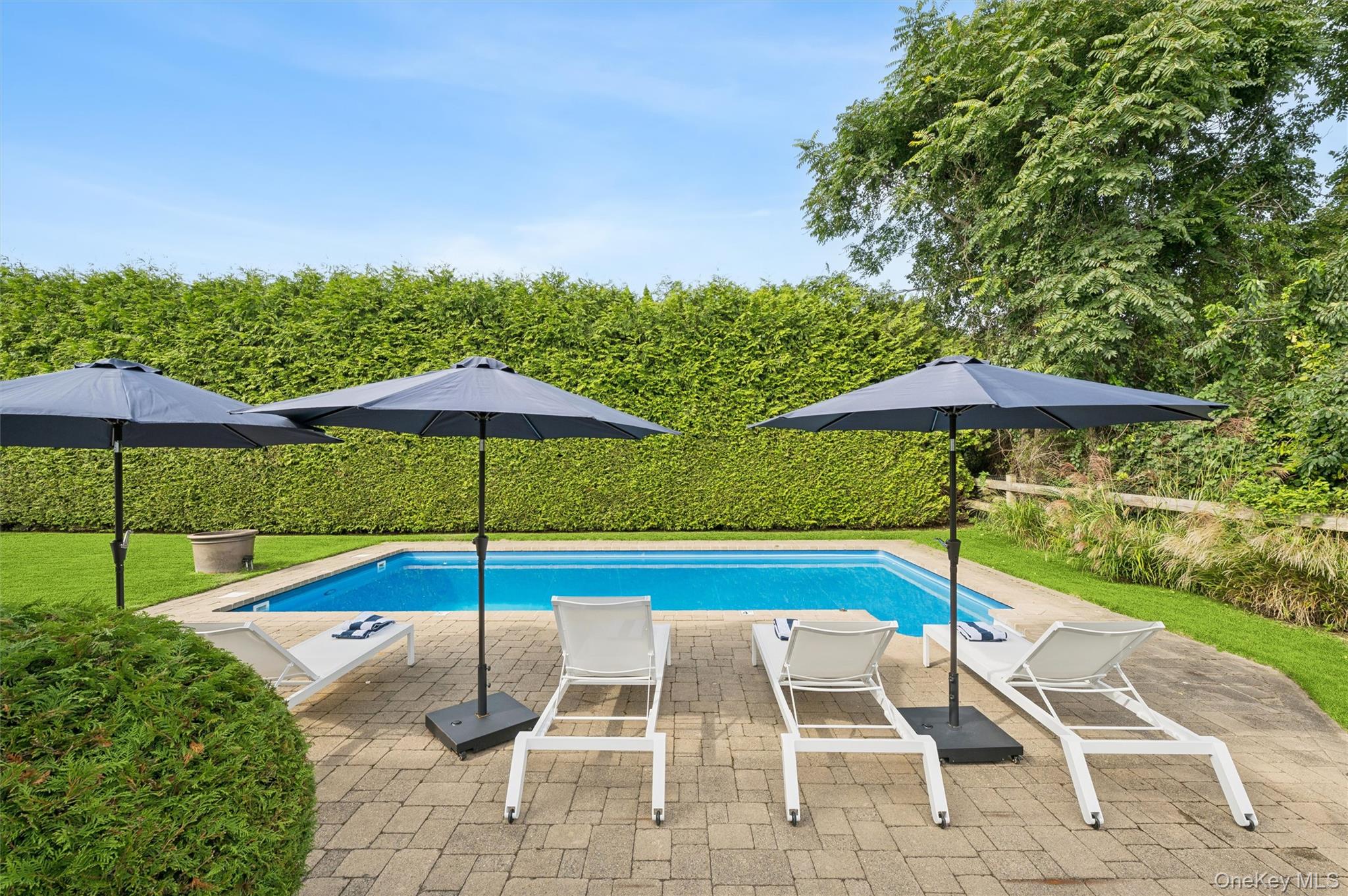 Outdoor pool with white lounge chairs and dark umbrellas on a paved patio, bordered by a tall hedge.