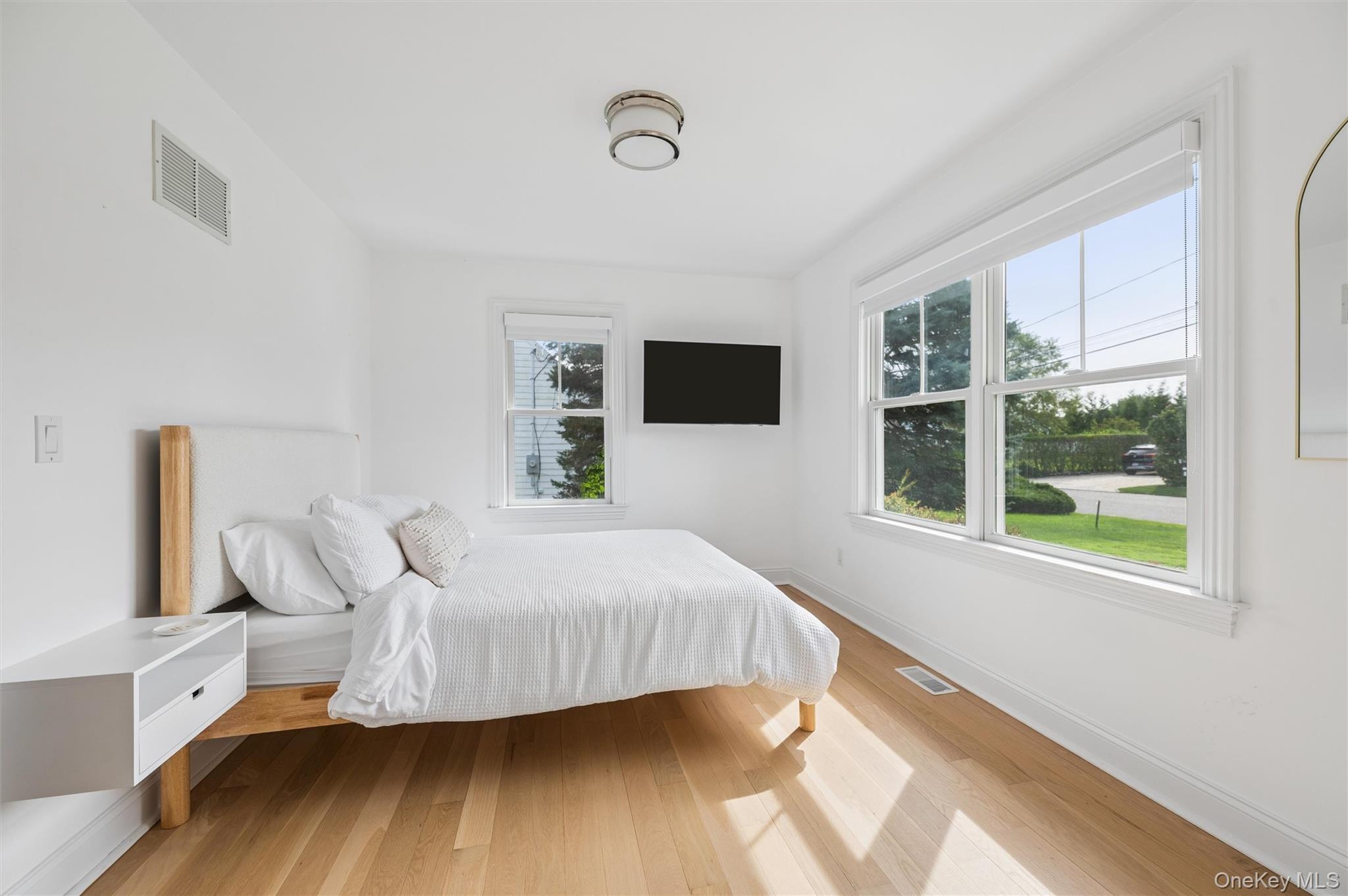 Bright bedroom with a white bed, light wood floors, and large windows.