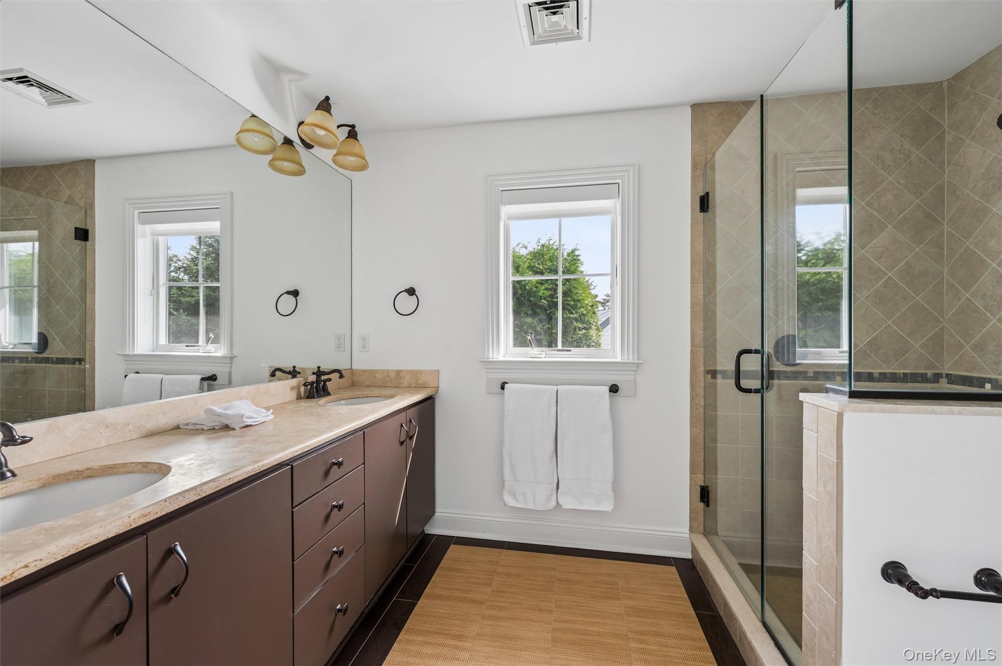 Bathroom with a long stone vanity, two windows, and a glass-enclosed tiled shower.