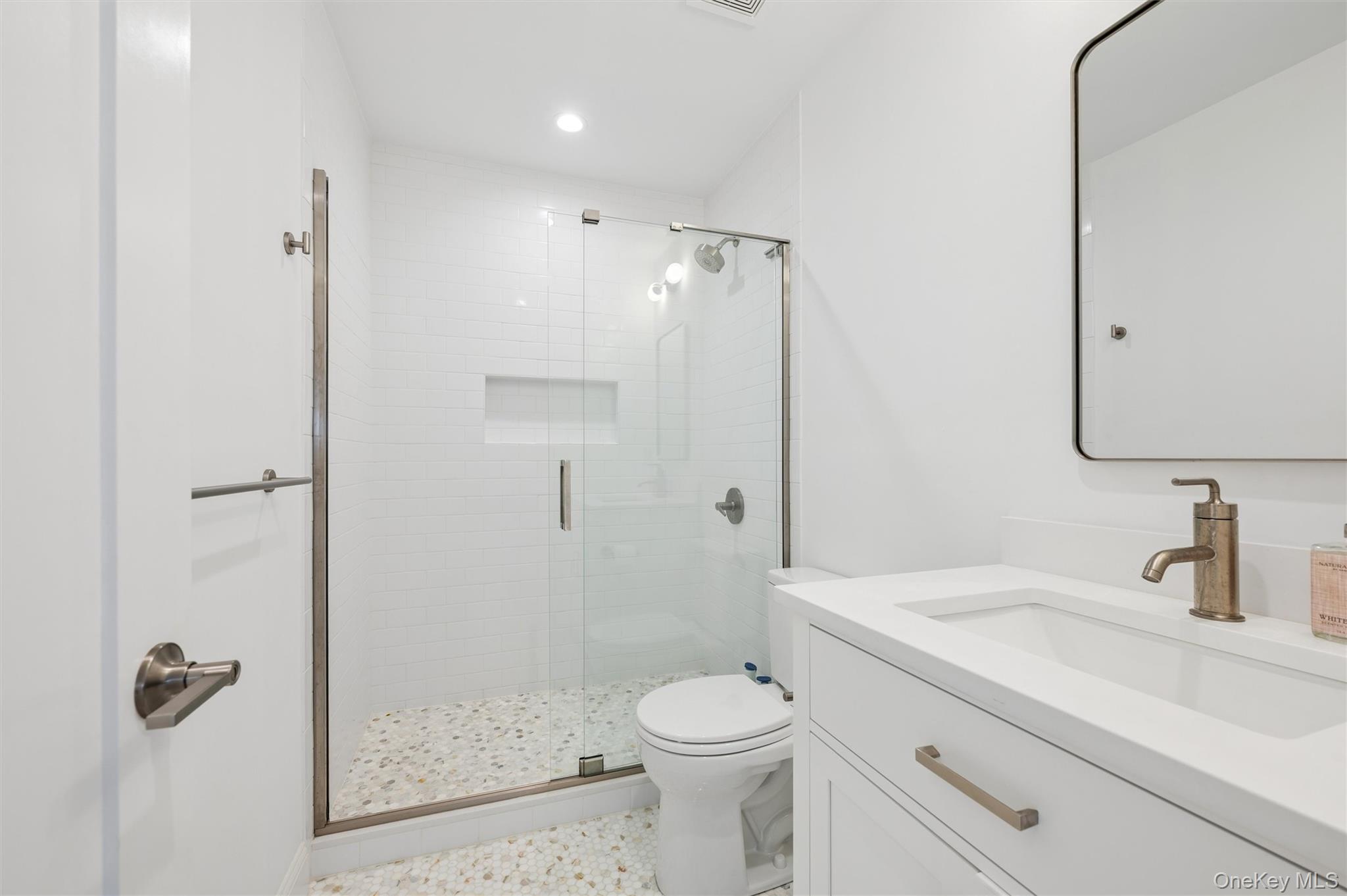White bathroom with a glass walk-in shower, pebble mosaic floor, and vanity with bronze fixtures.