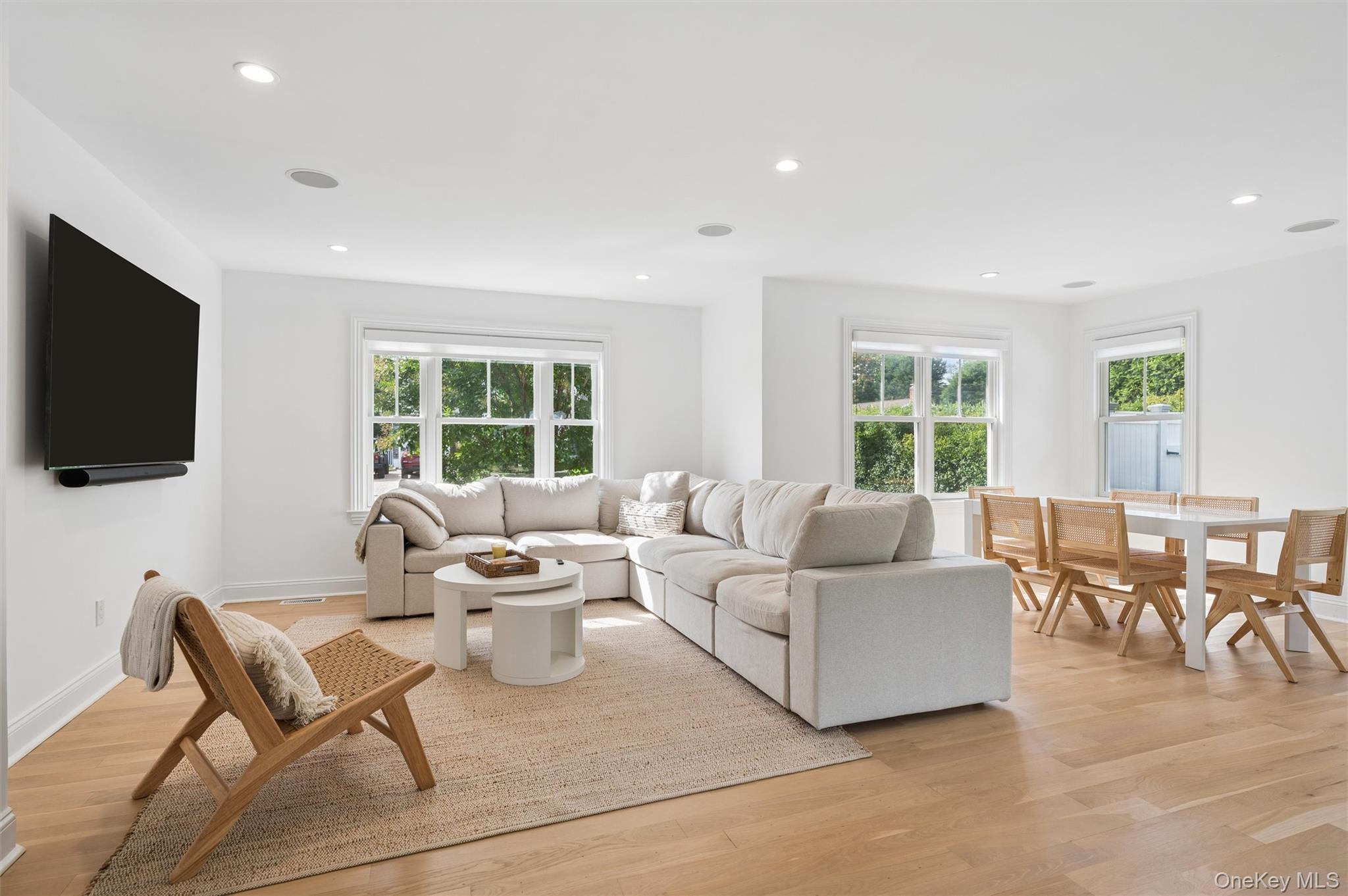 Open-plan living and dining room with a cream sectional, woven rug, and windows overlooking greenery.