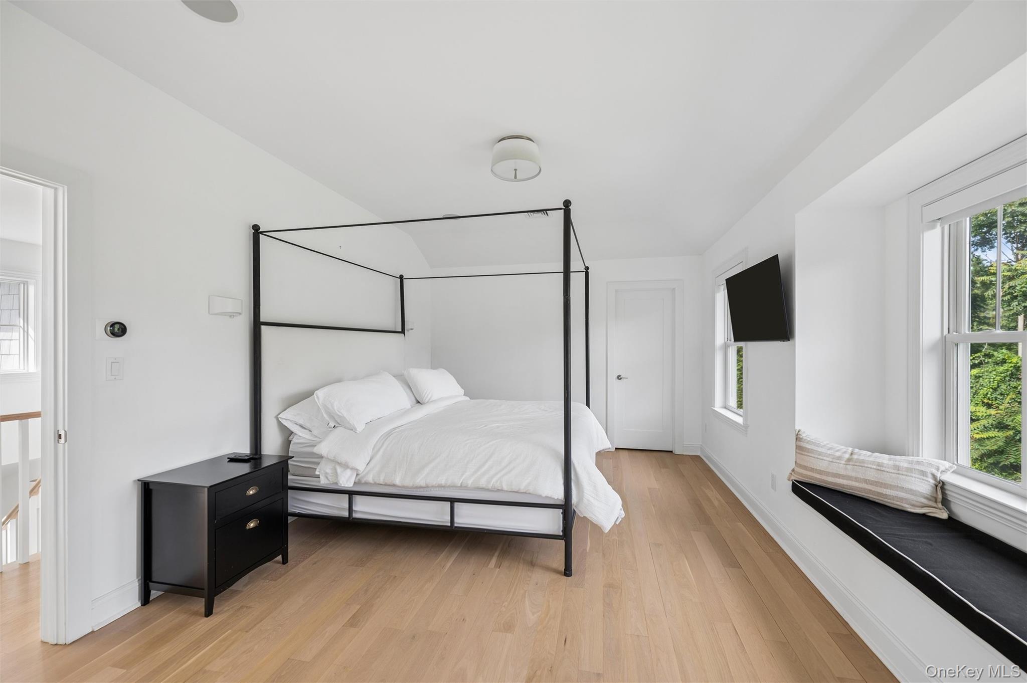 Bedroom with a black canopy bed, light wood floors, and a window seat overlooking greenery.