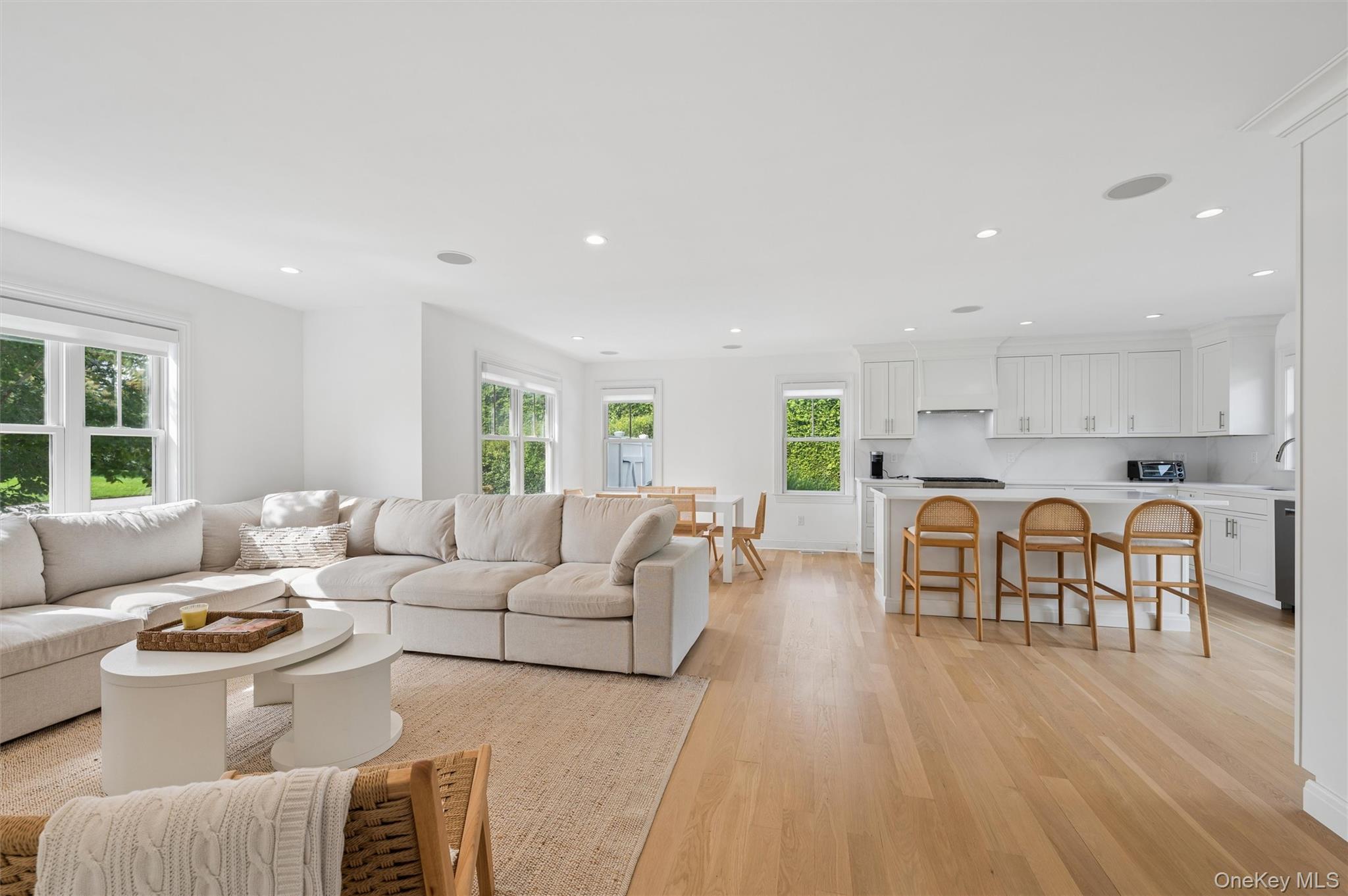 Open-plan living room and kitchen with a cream sectional, white cabinetry, and wooden island stools.