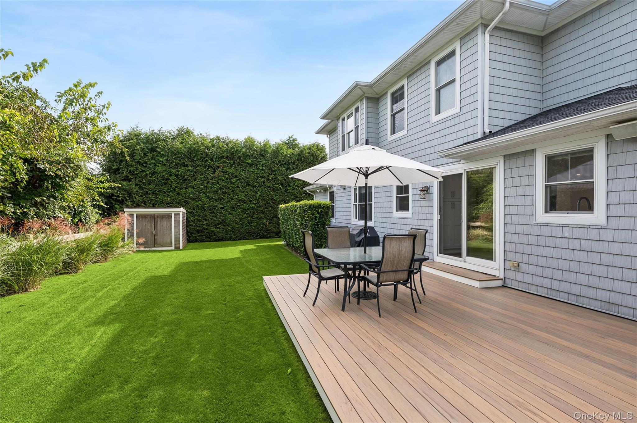 Gray-shingle home with a furnished deck, umbrella-shaded dining table, and hedge-lined lawn.