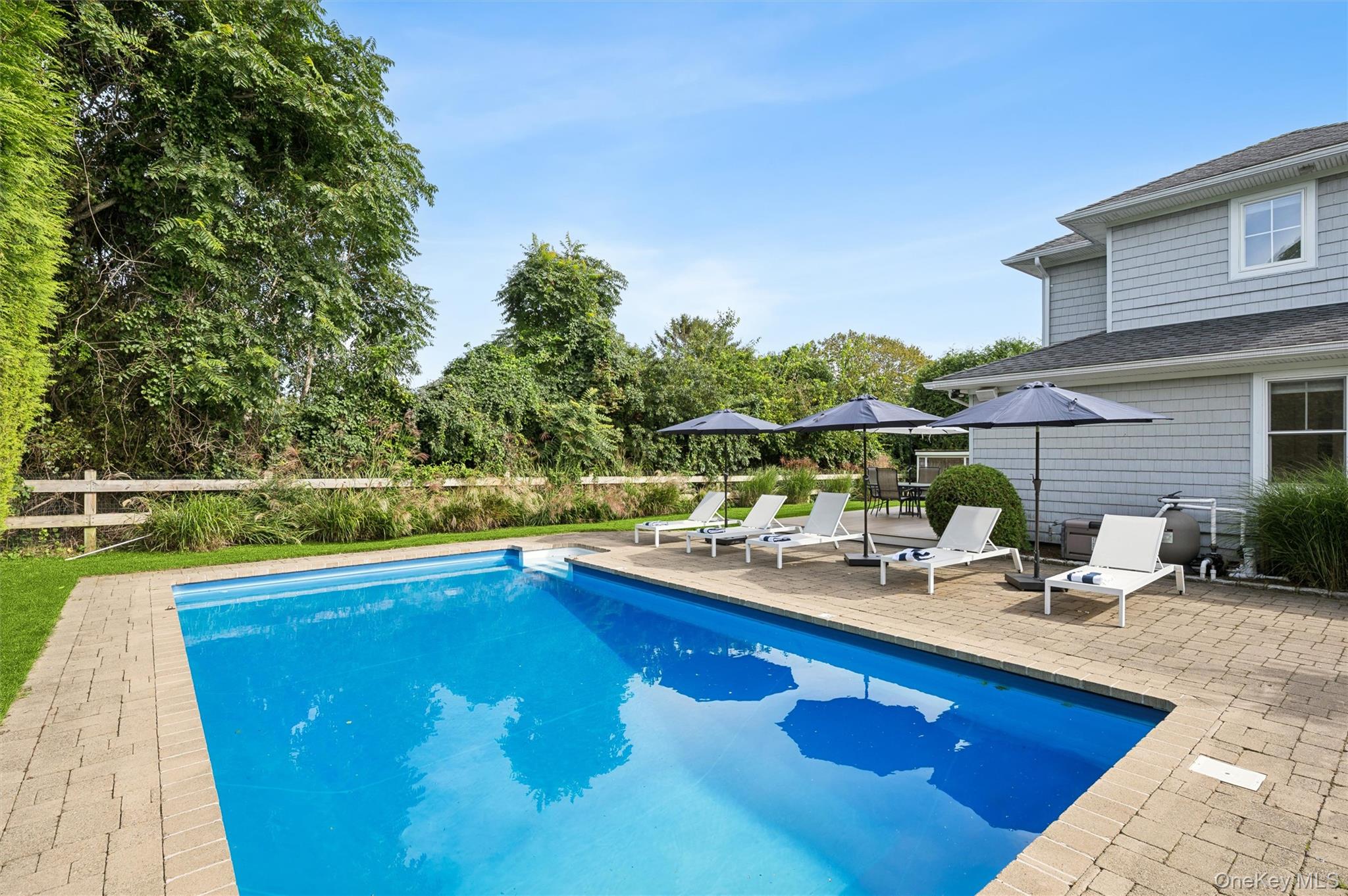 Backyard swimming pool with patio loungers and umbrellas beside a gray shingle-sided house.