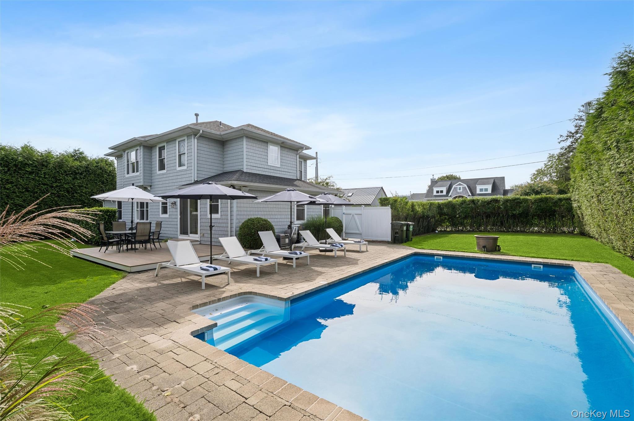 Gray two-story house beside a swimming pool, with a stone patio and shaded lounge chairs.