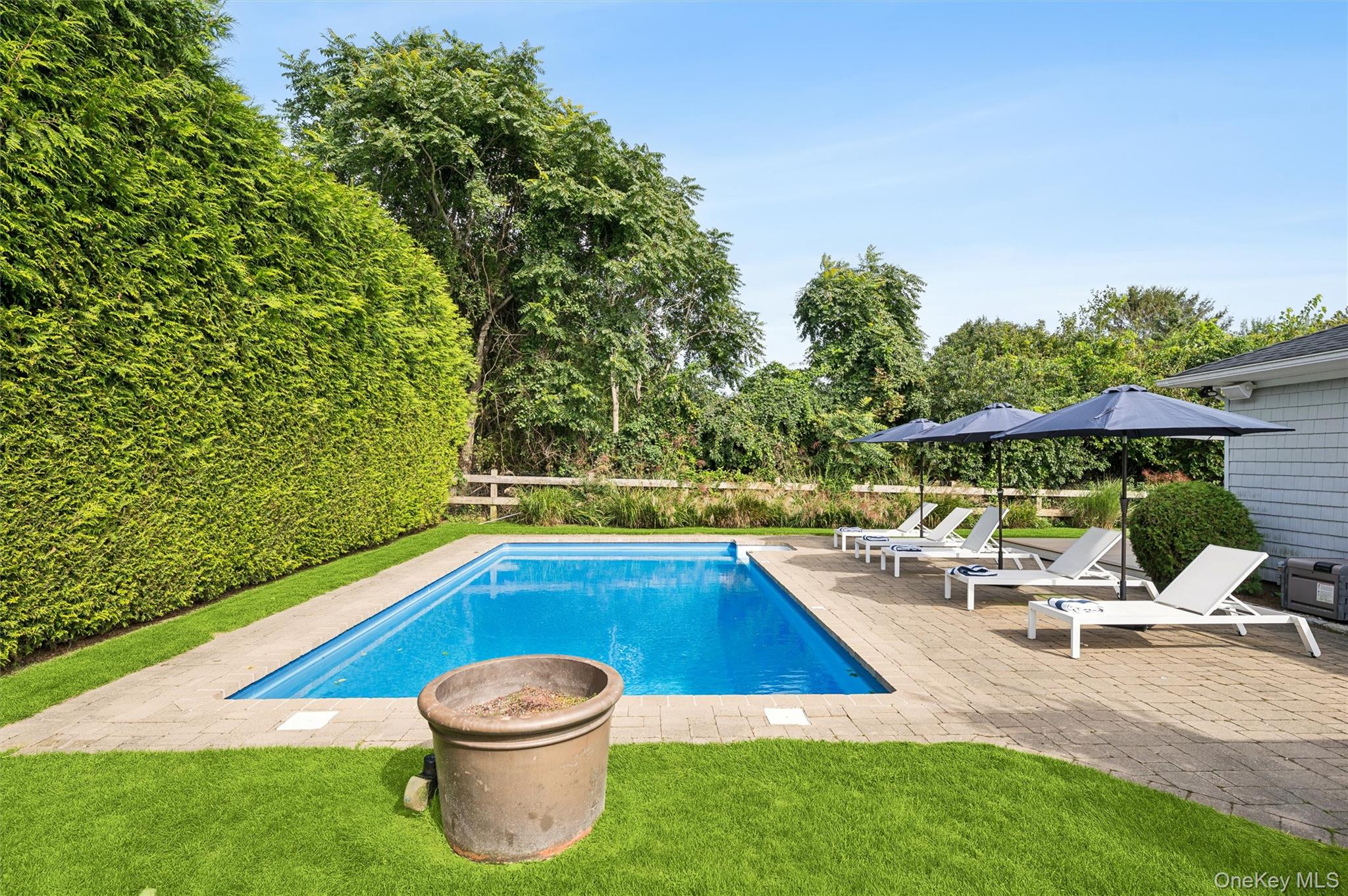 Backyard swimming pool beside a paver patio with white loungers and umbrellas.