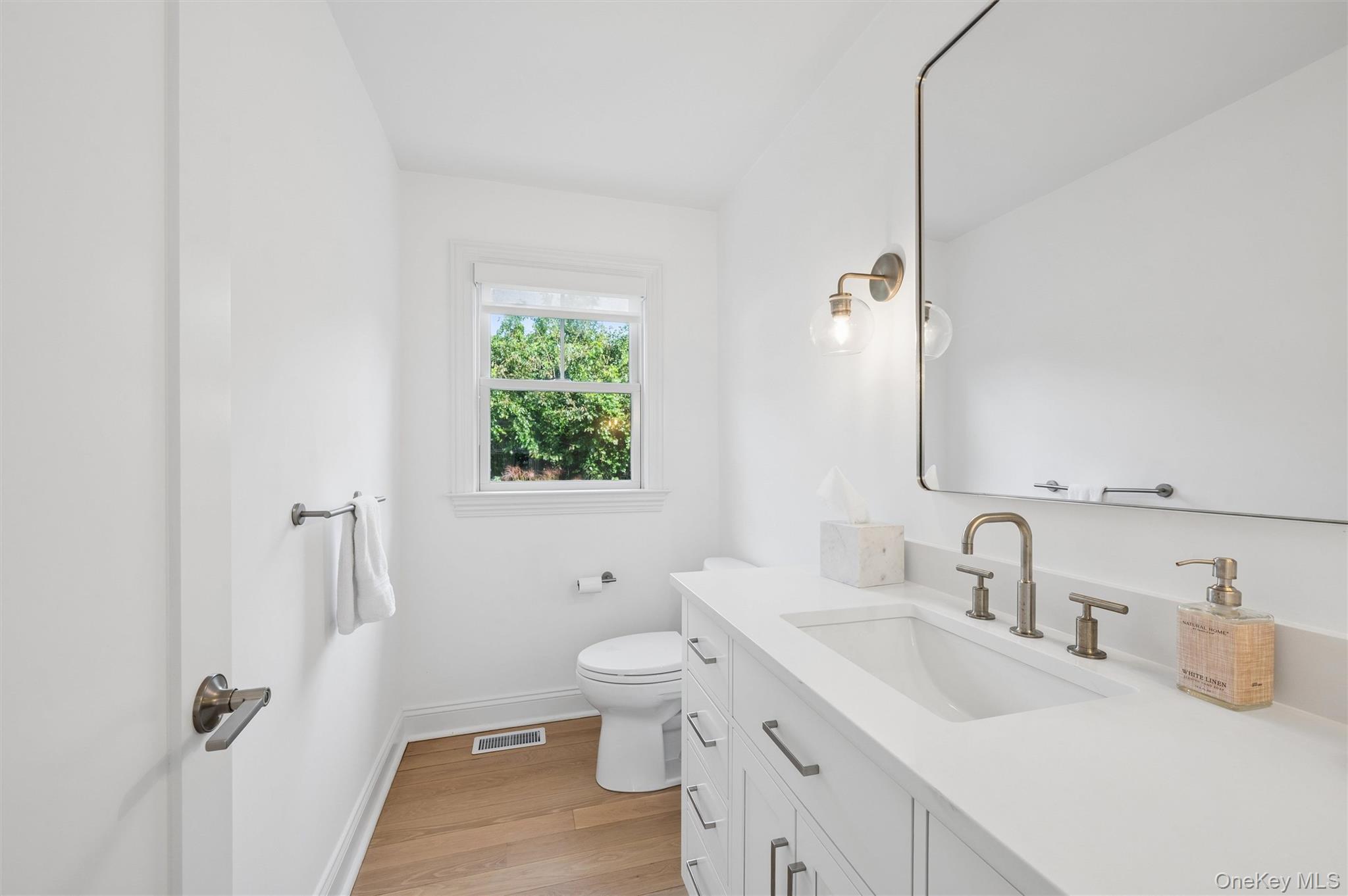 Bright white bathroom with a long vanity, brass fixtures, and a window overlooking greenery.