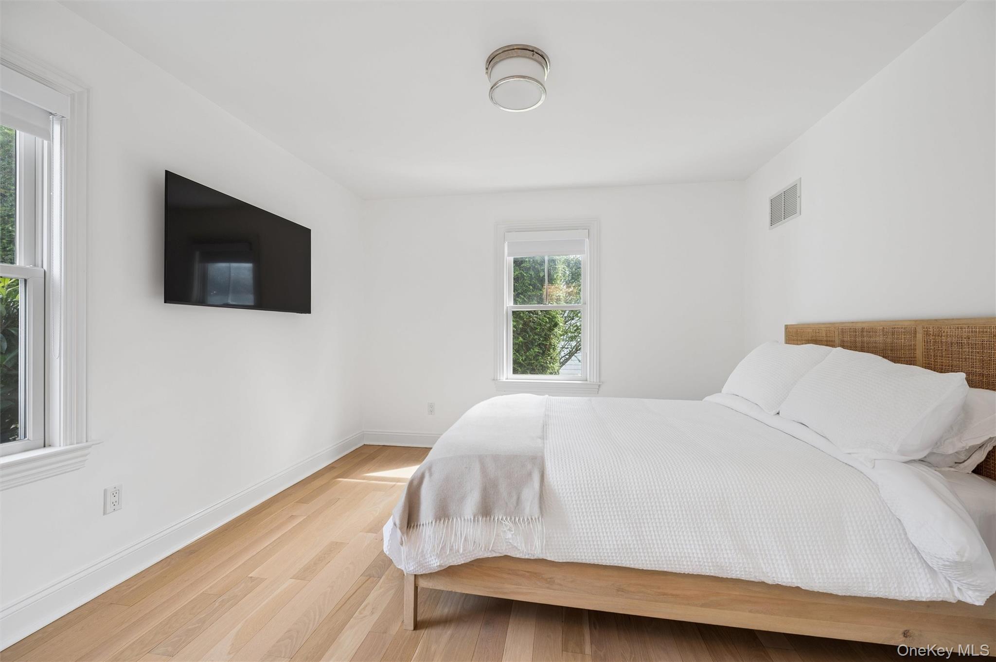 Bright bedroom with a large white bed, woven headboard, wall-mounted television, and light wood floors.