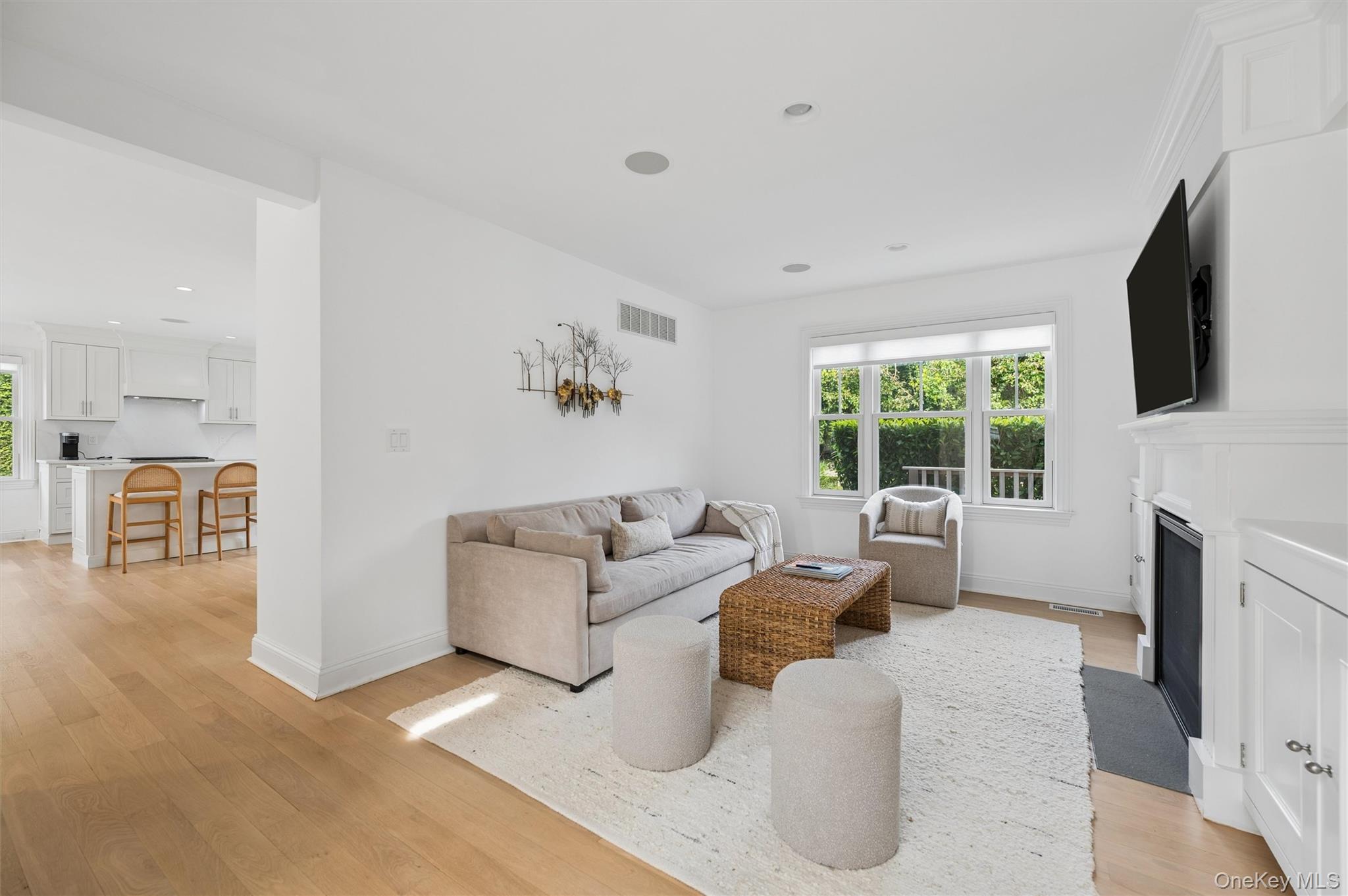 Living room with a white fireplace, neutral seating, large windows, and an adjoining kitchen.