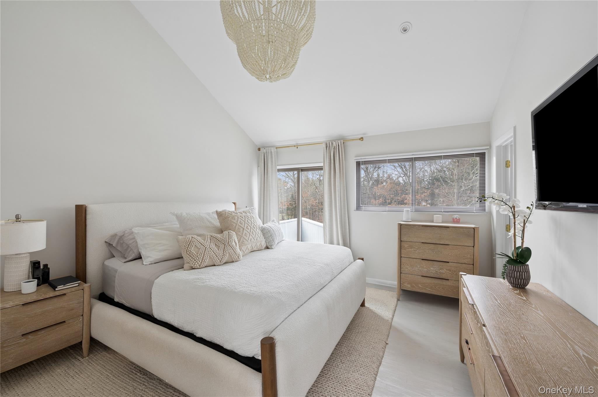 Bright bedroom with a white upholstered bed, wooden furniture, and windows overlooking bare trees.