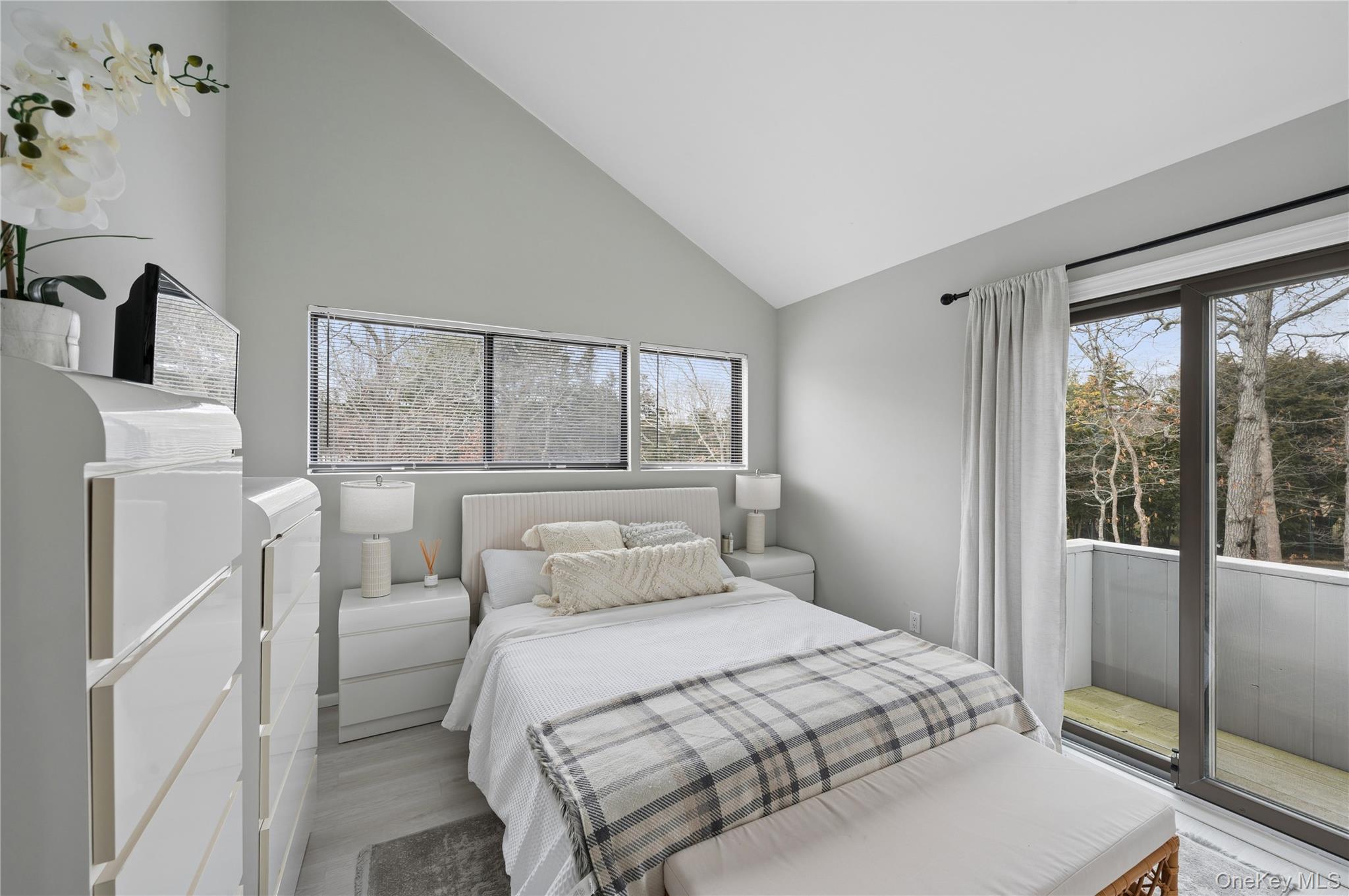 Staged bedroom with a vaulted ceiling, white bedding, and a sliding glass door to a tree-lined balcony.
