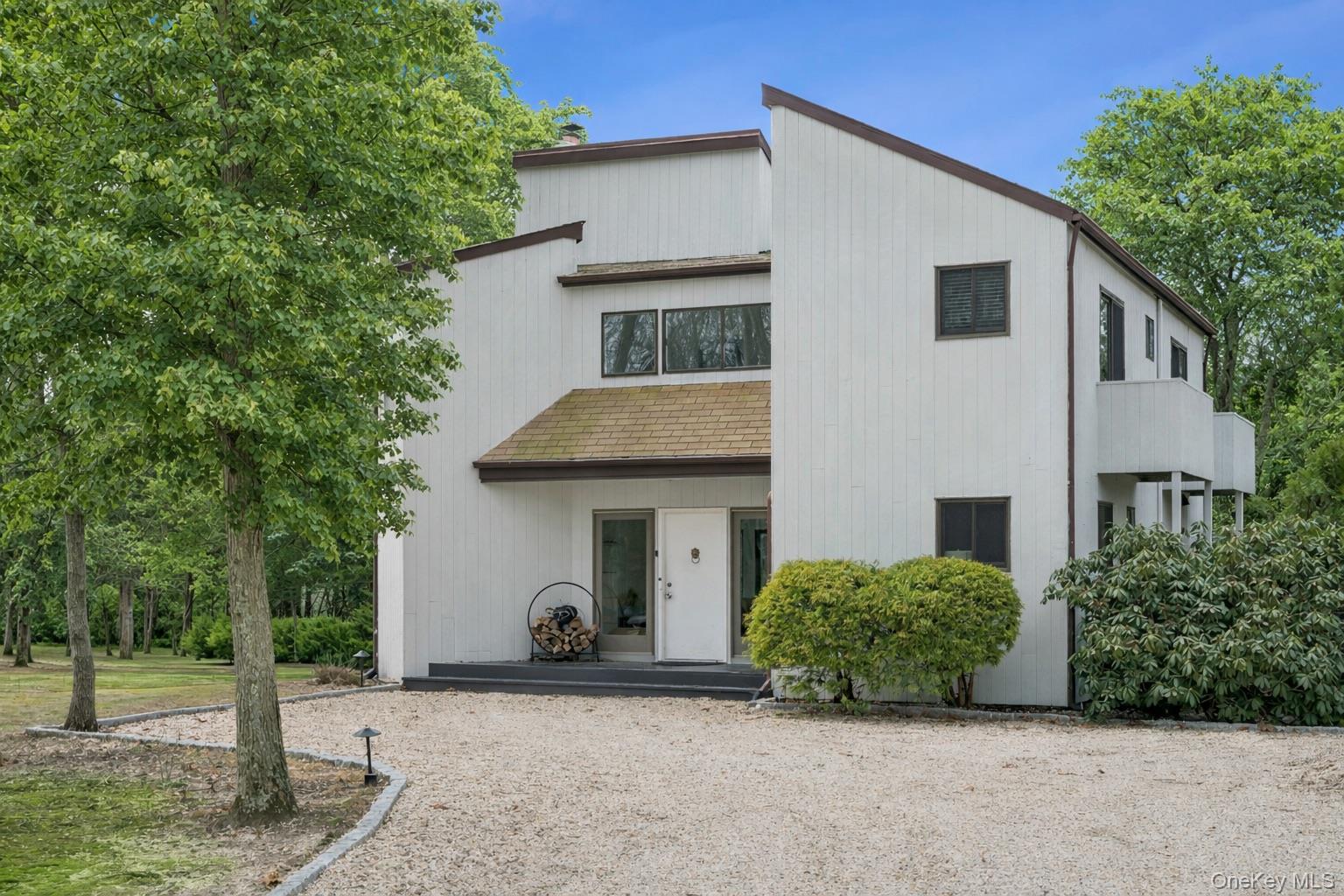White multi-level house with brown trim, set among leafy trees beside a gravel driveway.
