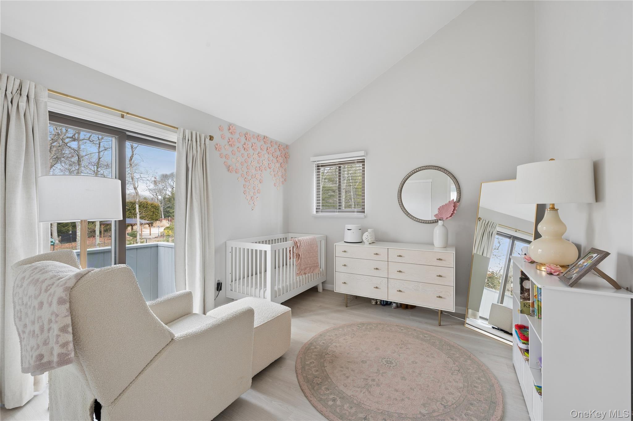 Nursery with a white crib, pink floral wall accents, and an upholstered armchair beside large windows.