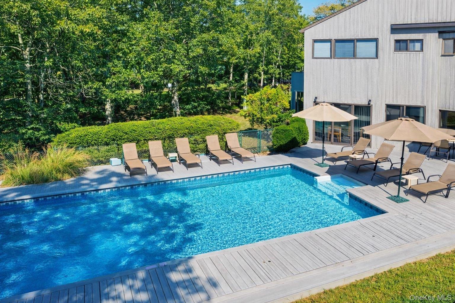 Outdoor swimming pool beside a modern gray home, with a wood deck, lounge chairs, and umbrellas.