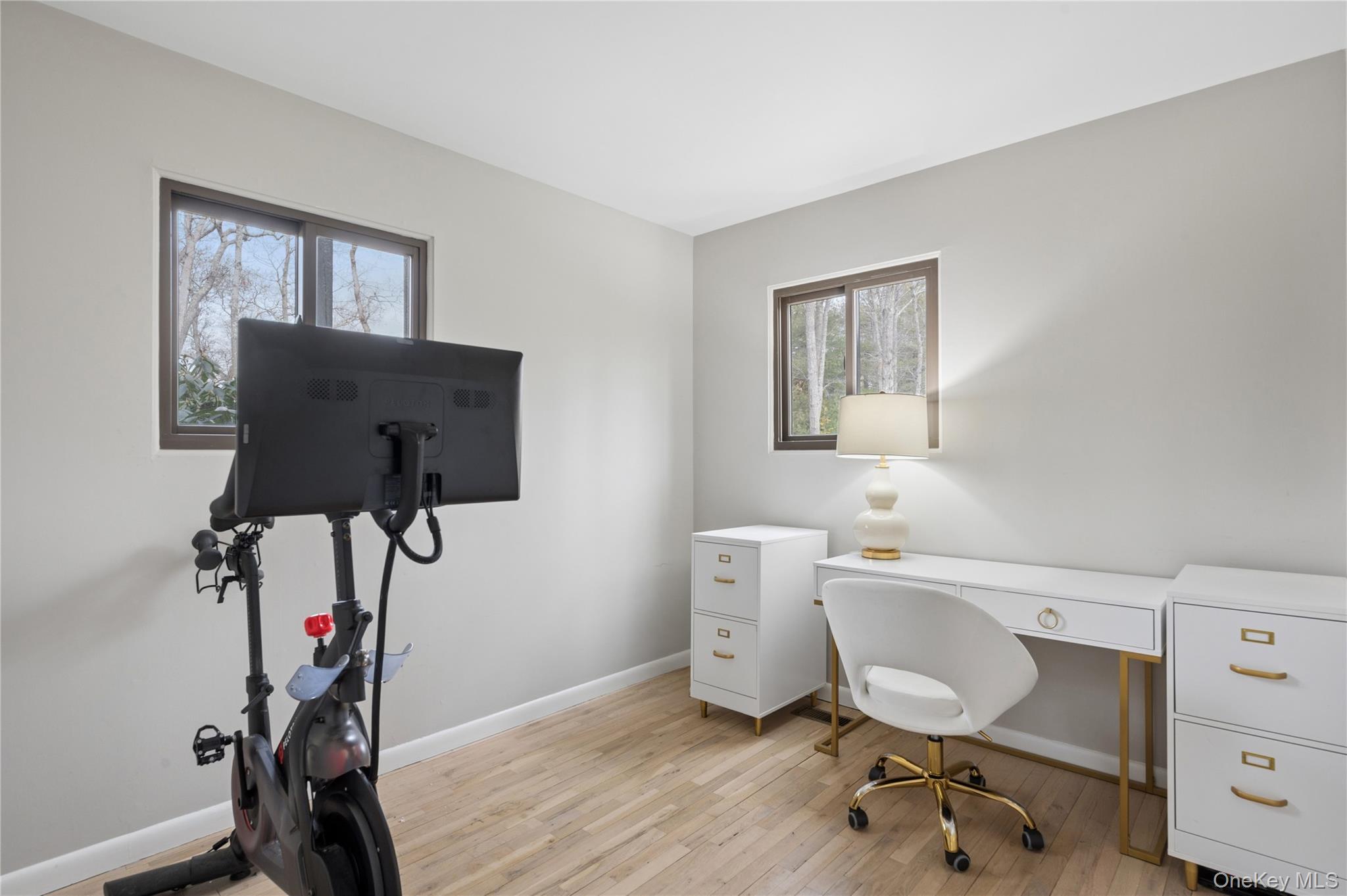 Home office with a white desk and gold accents beside a stationary exercise bike with a large monitor.